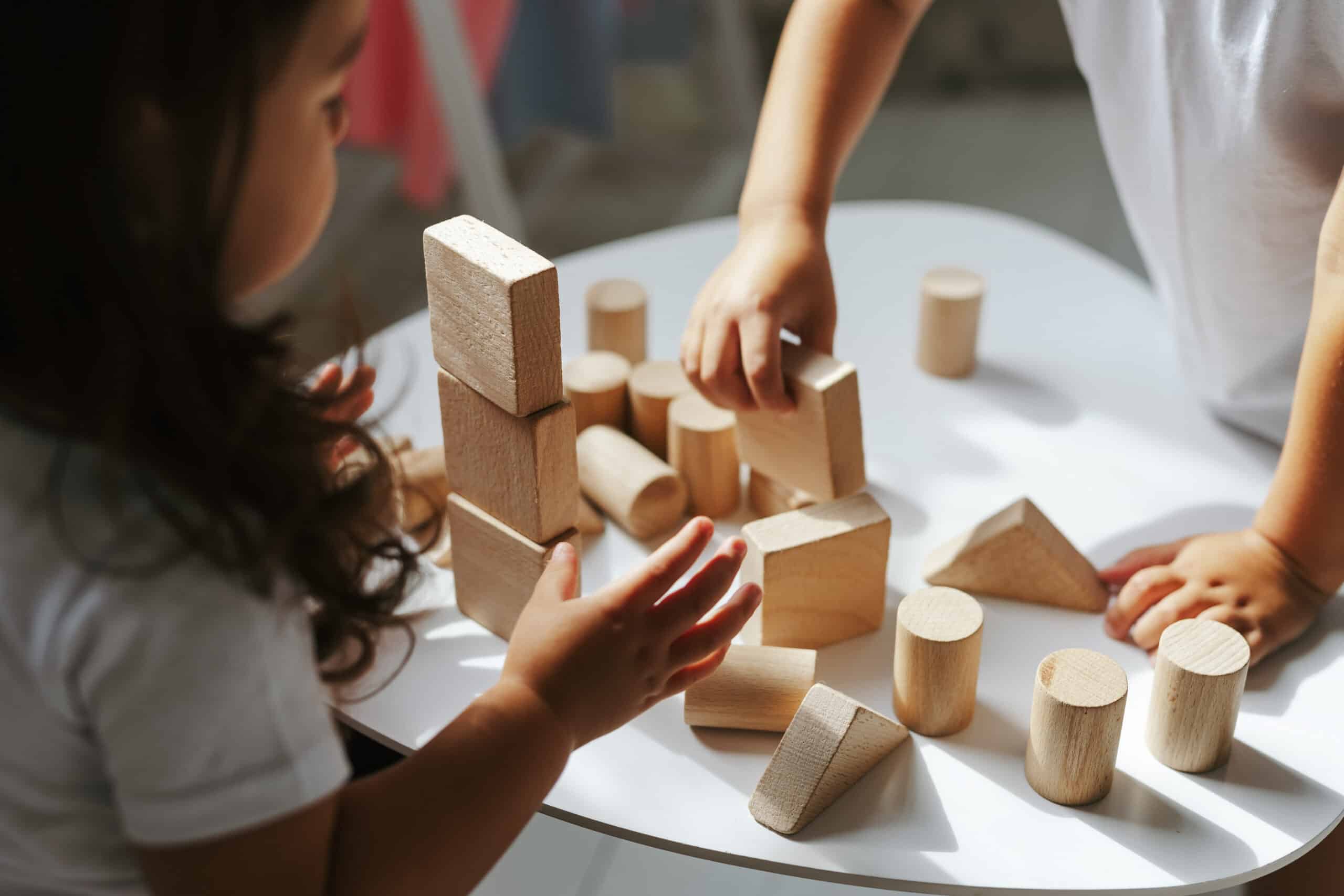 Deux enfants jouent ensemble avec des blocs de construction en bois clair empilés sur une table blanche, leurs mains sont visibles.
