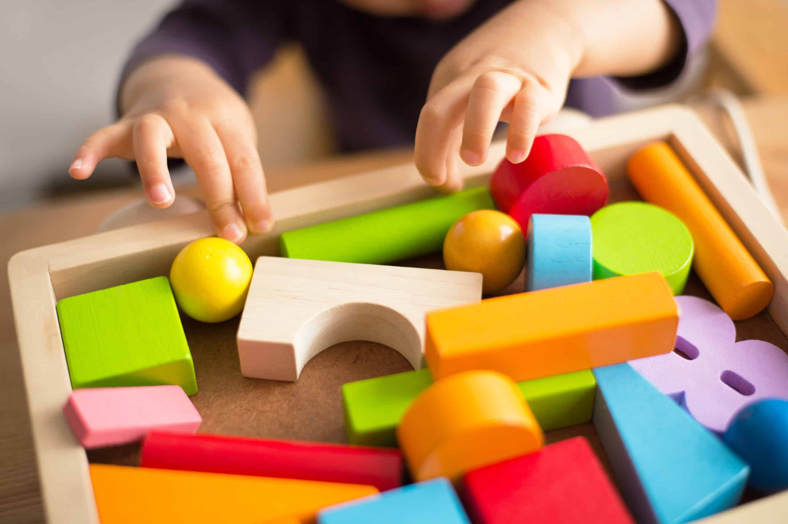 Mains d'enfant jouant avec des blocs de bois colorés (rond, cylindre, cube) dans un plateau en bois clair.