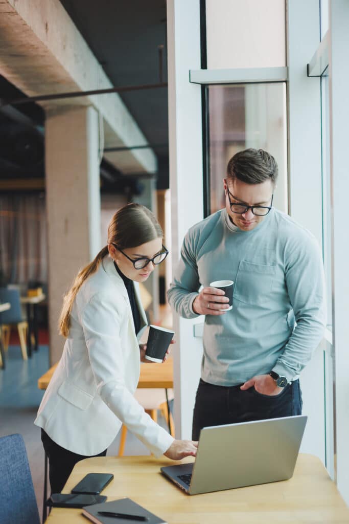 Collaboration de professionnels avec ordinateur et café Deux professionnels, un homme et une femme portant des lunettes, consultent un ordinateur portable sur un bureau, chacun tenant une tasse de café.