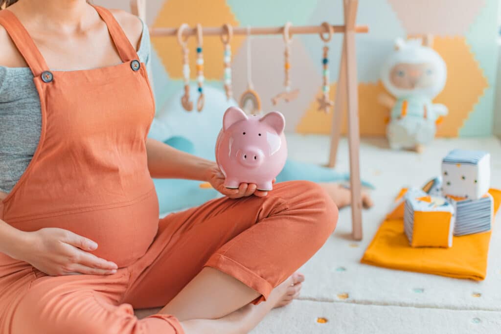 Épargne bébé : future maman prépare l'avenir Femme enceinte en salopette pêche tient une tirelire cochon rose près de son ventre, dans une chambre de bébé décorée.