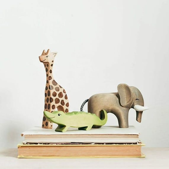 Figurines en bois d'animaux de la savane : girafe tachetée, alligator vert et éléphant gris, posées sur des livres.