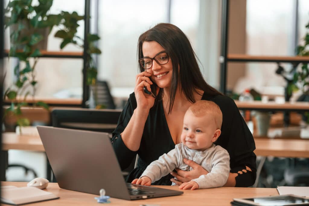 Une mère souriante, lunettes et haut noir, parle au téléphone en tenant son bébé blond devant un ordinateur portable au bureau.