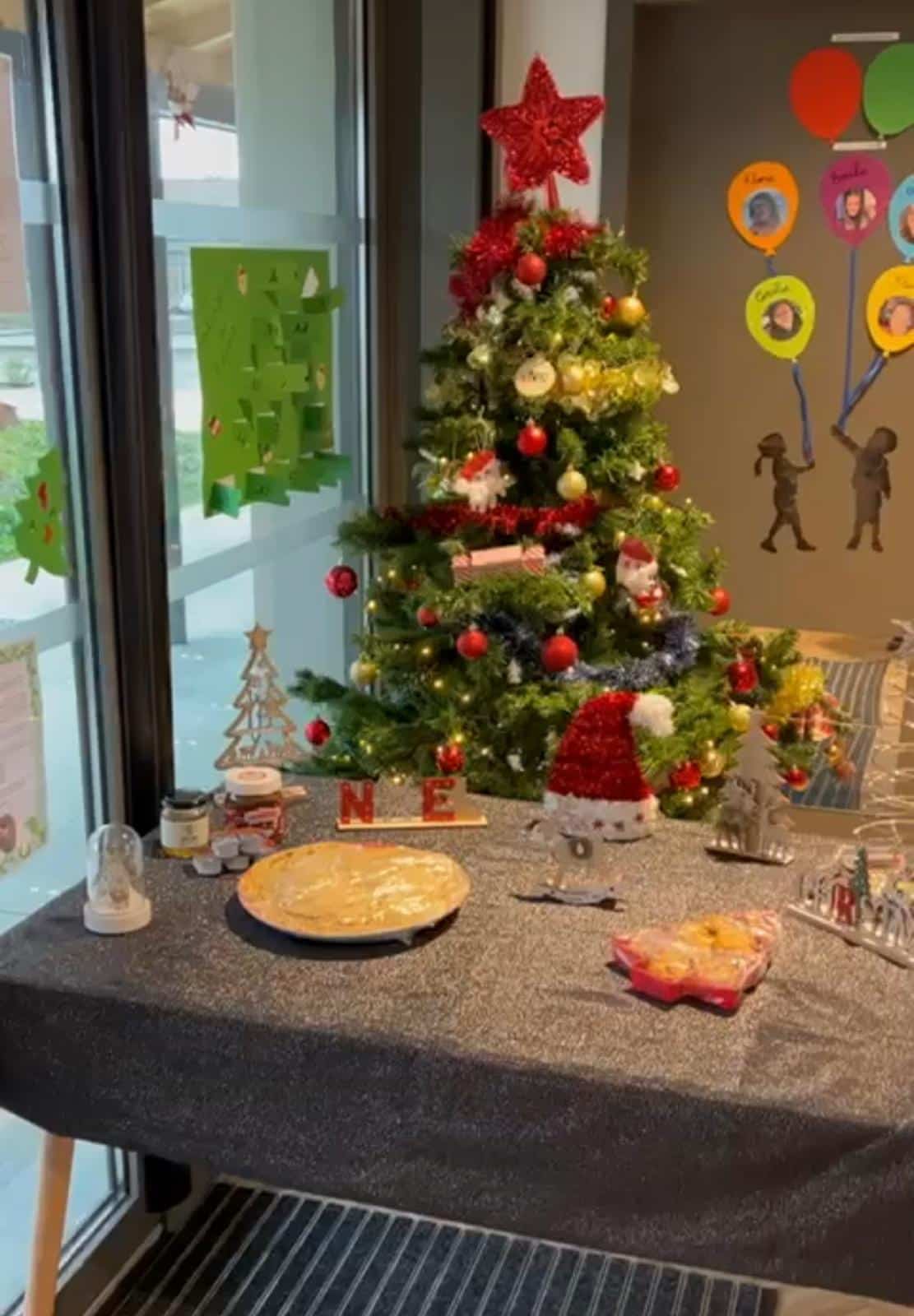 Table décorée pour Noël avec sapin, tarte, pot de Nutella, et décorations rouges et blanches. Des lettres formant "N E" sont visibles.