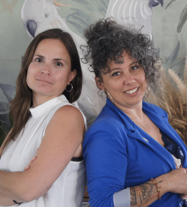 Partenaires souriantes posant devant un décor tropical Deux femmes souriantes, l'une en haut blanc et l'autre en blazer bleu, se tiennent dos à dos devant un mur décoré de perroquets et de feuillage.